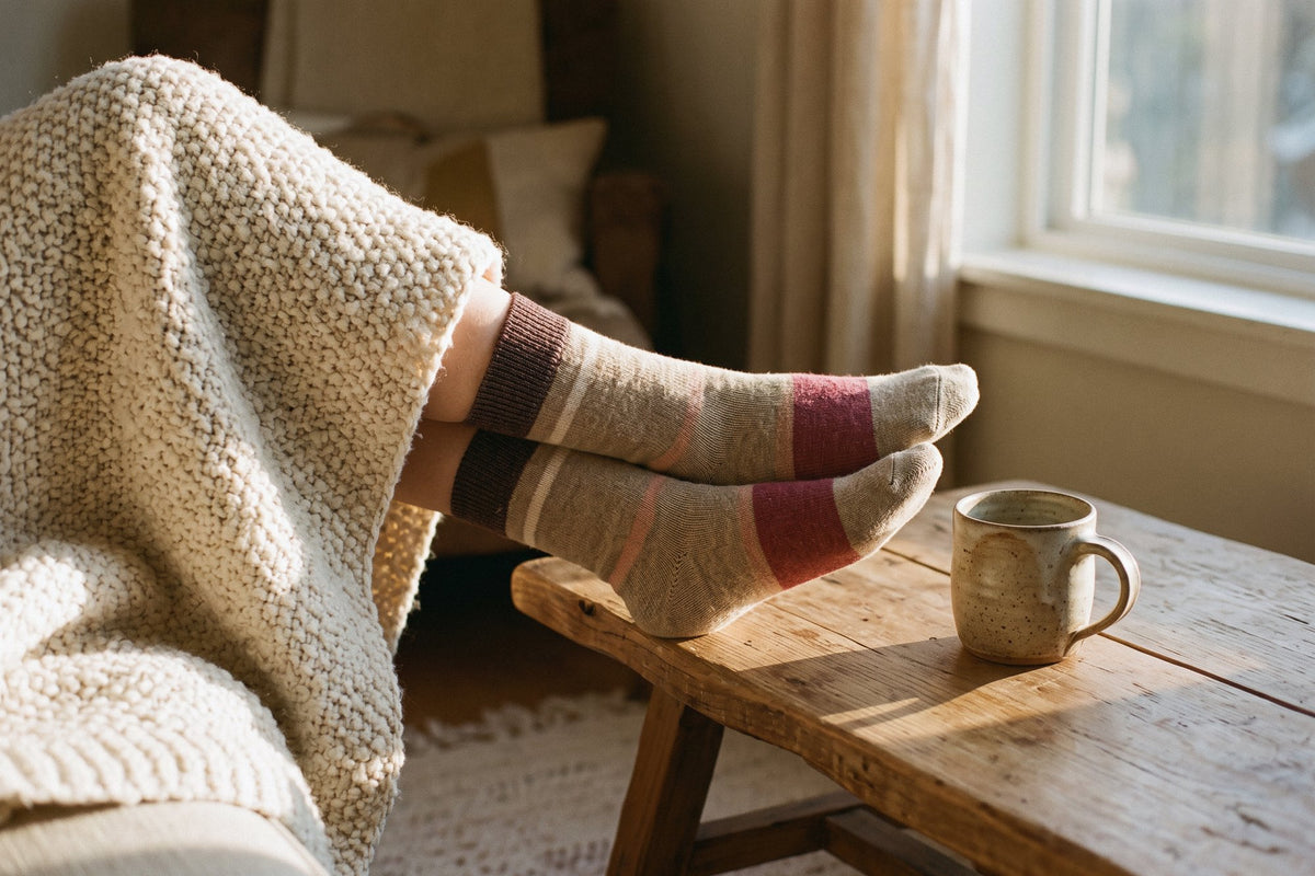 Calcetines de lana suave beige y malva puestos, pies sobre una mesa baja de madera junto a la ventana